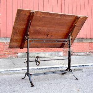 May include: An antique drafting table with a large, angled wooden surface. The table features a dark metal frame with adjustable height and angle mechanisms. The wooden surface is a warm brown color, showing signs of age and use.
