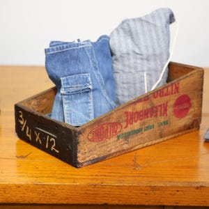 May include: An aged wooden crate with red and green text and a red circle contains folded denim clothing. The crate is on a wooden surface, with additional folded denim items nearby. The crate has the dimensions 3/4 x 12 written on the side.