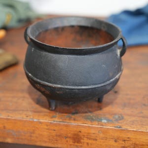 May include: A black cast iron cauldron with a rounded body, a wide open top, and two small side handles. The vessel features three short legs at the base and shows signs of rust and wear on the interior and exterior surfaces of the metal.