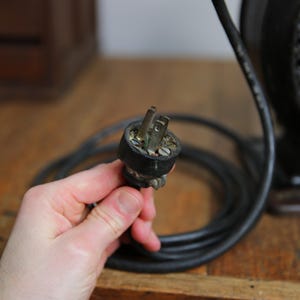 May include: Close-up of a black electrical cord with a two-prong plug. The plug is connected to a vintage electrical device.