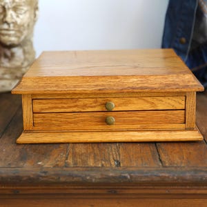 May include: A small, two-drawer wooden box with a light brown finish. The box has brass knobs on the drawers and is sitting on a wooden surface. The box is likely used for storing small items.