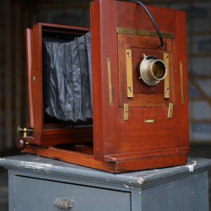 May include: A vintage wooden camera with a dark brown finish, featuring a bellows and brass accents. The camera is mounted on a gray cabinet. The lens is visible, and the camera appears to be in good condition.