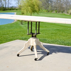 May include: A white wooden table with a long rectangular top and a four-legged base with wheels. The table is on a concrete surface with green grass in the background.