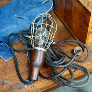 May include: An antique work light with a metal cage, a wooden handle, and a black cord. The light bulb is visible inside the cage. The light is resting on a wooden surface with a pair of blue jeans.