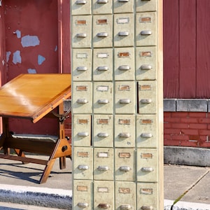 May include: A tall, vintage, pale green metal cabinet with 30 drawers. Each drawer has a metal pull handle and a small, rectangular label holder. The cabinet is standing on a concrete surface.