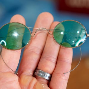 May include: Oval-shaped spectacles featuring dark green tinted lenses and a thin, metallic wire frame. The glasses include a delicate bridge and thin, curved wire temples that extend downward, designed to rest securely around the ears.