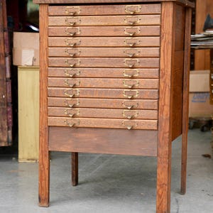 May include: A tall, rectangular wooden cabinet featuring ten horizontal drawers stacked vertically. Each drawer is equipped with a brass label holder and a small pull handle. The cabinet is constructed from brown wood with a visible grain pattern.