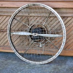 May include: A vintage, silver-colored, propeller fan with a wire mesh cage. The fan is mounted on a wooden wall.