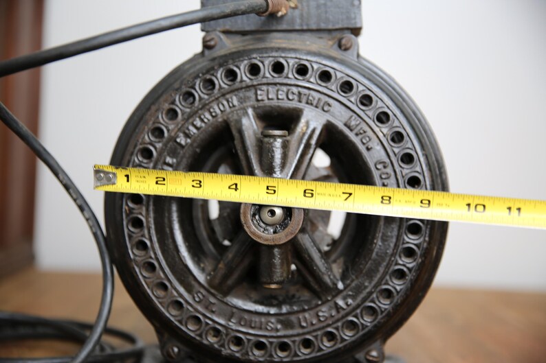 May include: A black cast iron Emerson Electric Mfg. Co. motor with a 11-inch diameter. The motor has a circular, black, metal casing with a series of holes around the edge. The motor is labeled "Emerson Electric Mfg. Co. St. Louis, U.S.A."
