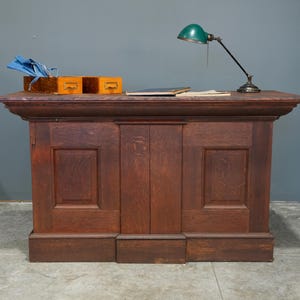 May include: A dark wood desk with a green desk lamp, small wooden boxes, and a book. The desk has a rectangular shape with a decorative edge and paneling on the front. The lamp has a green shade and a black arm.