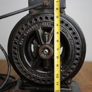 May include: A black cast iron electric motor with a 12-inch diameter. The motor has a round, black metal casing with a series of holes around the edge. The motor is labeled "The Emerson Electric Mfg. Co. St. Louis, Mo."