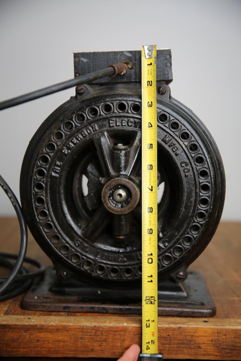 May include: A black cast iron electric motor with a 12-inch diameter. The motor has a round, black metal casing with a series of holes around the edge. The motor is labeled "The Emerson Electric Mfg. Co. St. Louis, Mo."