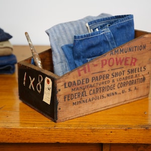 May include: An antique wooden box with the text "HI-POWER" and "LOADED PAPER SHOT SHELLS" contains folded denim clothing. A stack of folded denim shirts and a paintbrush are also visible on a wooden surface.