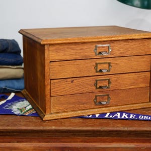 May include: A vintage wooden storage cabinet with three drawers, each featuring a metal label holder. The cabinet is made of light brown wood and sits on a wooden surface. A blue pennant with white text is visible.