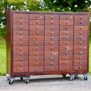 May include: A large, rectangular wooden cabinet featuring forty individual drawers arranged in four vertical columns. Each drawer is equipped with a small round wooden knob and a rectangular metal label holder. The unit is mounted on four swivel casters.