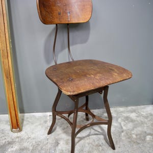 May include: An antique wooden chair with a curved back and seat, supported by a dark metal frame. The chair has a rustic, worn appearance, suggesting it is vintage or reclaimed. The design is simple and functional, suitable for various interior styles.