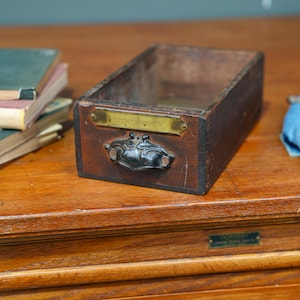 May include: A vintage wooden drawer with a dark brown finish. It features a brass plate and a decorative metal handle. The drawer is empty and sits on a wooden surface, suggesting its use for storage or organization.