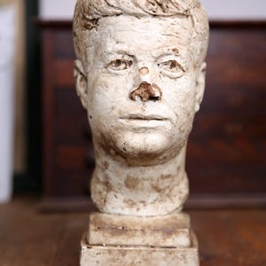 May include: A white plaster bust of a man with short hair. The bust is on a white pedestal. The man's face is weathered and has a chipped nose.