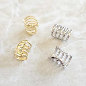 May include: Four ear cuffs in gold and silver tones. Two gold ear cuffs have a spiral design, and the other two are silver. One silver ear cuff has a coiled design, and the other has a linear design. The ear cuffs are on a white marble surface.