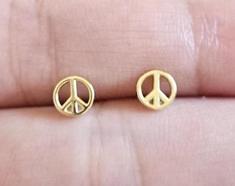 May include: A pair of small, gold-colored peace sign stud earrings. The earrings are round with the iconic peace symbol in the center. The earrings are displayed against a skin-toned background.