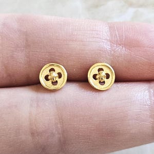 May include: A pair of small, gold-colored button earrings. Each earring has a circular shape with a four-petal design in the center. The earrings are shown against a soft, neutral background.