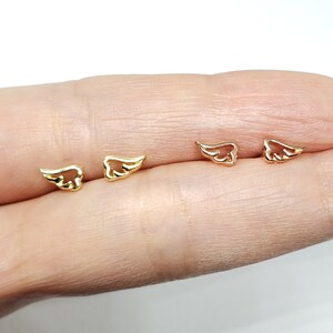May include: Four pairs of small gold and rose gold angel wing earrings. The earrings are displayed on a finger.