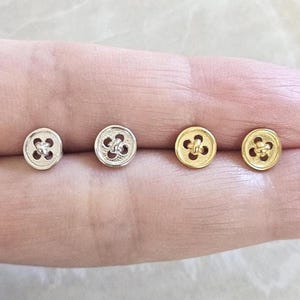 May include: Four button-shaped stud earrings. Two are silver-colored, and two are gold-colored. Each earring has a four-petal design in the center, resembling a button's stitching holes. The earrings are small and delicate, suitable for everyday wear.