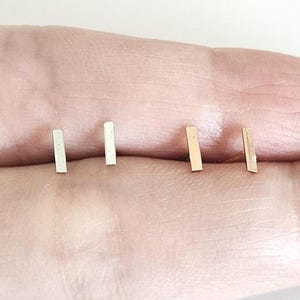 May include: A close-up of four small, rectangular bar earrings. Two are silver-colored, and two are gold-colored. The earrings are displayed on a light skin tone background.
