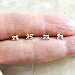 May include: Four tiny bow earrings in gold and silver. The earrings are shown on a finger.