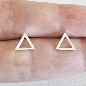 May include: A pair of small, open triangle stud earrings. The earrings are silver-colored and have a simple, geometric design. The earrings are shown on a light-colored background.