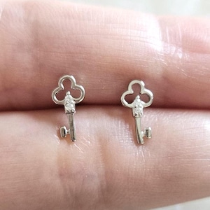 Puede incluir: Un par de pendientes de bot&oacute;n plateados con forma de llave. Cada pendiente presenta un dise&ntilde;o de llave con una parte superior de tres l&oacute;bulos y una peque&ntilde;a gema transparente en el centro de la cabeza de la llave. Los pendientes son peque&ntilde;os y delicados.