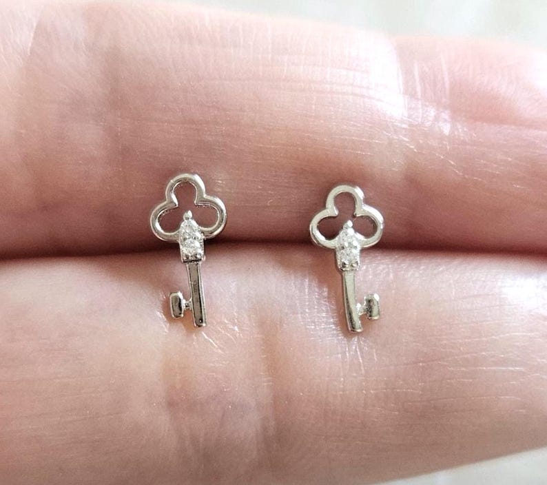Puede incluir: Un par de pendientes de bot&oacute;n plateados con forma de llave. Cada pendiente presenta un dise&ntilde;o de llave con una parte superior de tres l&oacute;bulos y una peque&ntilde;a gema transparente en el centro de la cabeza de la llave. Los pendientes son peque&ntilde;os y delicados.