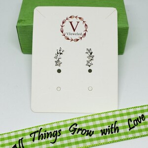 May include: A pair of silver star earrings on a white card with the text "VJeweled" and a floral design. The card is on top of a green box. A green and white gingham ribbon with the text "All Things Grow with Love" is in the foreground.