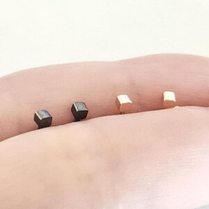 May include: Four small, cube-shaped stud earrings in varying metallic finishes. Two are dark, possibly black, and two are a light gold color. The earrings are displayed on a light skin tone background.