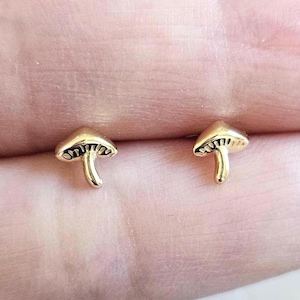 May include: A pair of small, gold-colored mushroom stud earrings. The earrings have a detailed cap and stem design, showcasing a whimsical and nature-inspired aesthetic. The earrings are presented on a soft, neutral background.