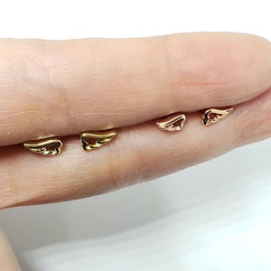May include: Three pairs of gold angel wing earrings. The earrings are shown on a finger.