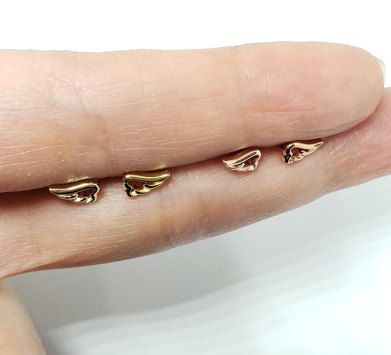 May include: Three pairs of gold angel wing earrings. The earrings are shown on a finger.