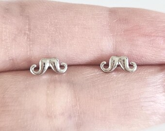 May include: A pair of small, silver-toned stud earrings shaped like stylized mustaches. Each earring features a smooth, reflective metallic finish with curved, upturned ends, designed as a symmetrical set of two individual mustache-shaped pieces.