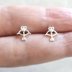 May include: A pair of silver stud earrings featuring a small bow and arrow design with a clear crystal heart in the center.