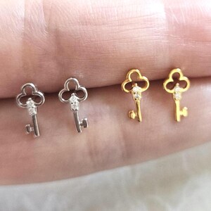 Puede incluir: Dos pares de pendientes con forma de llave, uno plateado y otro dorado. Cada llave tiene una parte superior decorativa y una peque&ntilde;a gema transparente. Los pendientes se muestran sobre una superficie clara.