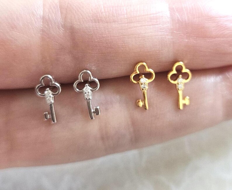 Puede incluir: Dos pares de pendientes con forma de llave, uno plateado y otro dorado. Cada llave tiene una parte superior decorativa y una peque&ntilde;a gema transparente. Los pendientes se muestran sobre una superficie clara.