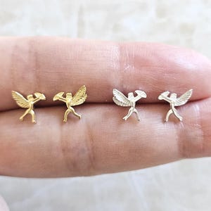 May include: Four small angel-shaped stud earrings. Two pairs of earrings are visible, with one pair in gold and the other in silver. The angels have outstretched wings and are holding a small object. The earrings are displayed on a light-colored surface.