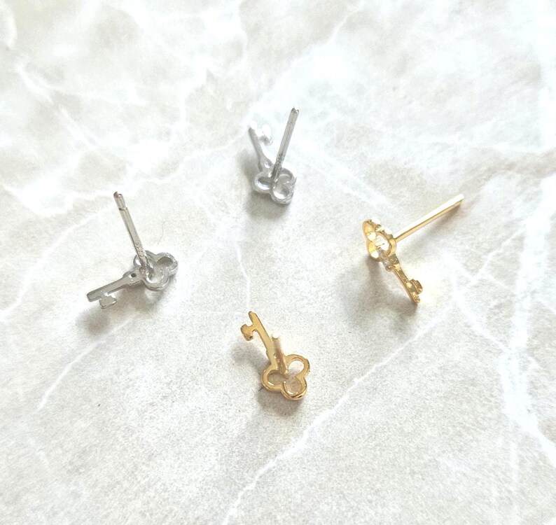 Puede incluir: Cuatro pendientes con forma de llave en tonos plateados y dorados est&aacute;n dispersos sobre una superficie clara. Los pendientes presentan dise&ntilde;os de ojo de cerradura y lazo, mostrando una delicada est&eacute;tica de joyer&iacute;a.