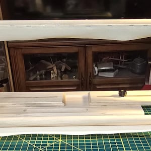 May include: A wooden embroidery hoop stand with a light-colored wood finish. The stand features a rectangular base, adjustable side supports, and a top bar to hold the fabric. The stand is designed for needlework projects.