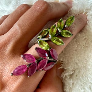 Może przedstawiać: Dwa srebrne pierścionki w kształcie liści, jeden z zielonymi kamieniami szlachetnymi, a drugi z różowymi kamieniami szlachetnymi. Pierścionki zdobione są wieloma owalnymi kamieniami ułożonymi tak, aby przypominały liście. Pierścionki są prezentowane na dłoni.