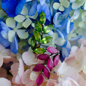 Pu&ograve; includere: Orecchini a forma di foglia con gemme verdi e magenta. Sono presentati su uno sfondo di fiori di ortensia blu, bianchi e rosa. Le gemme sono tagliate a forma ovale e disposte a forma di foglie.
