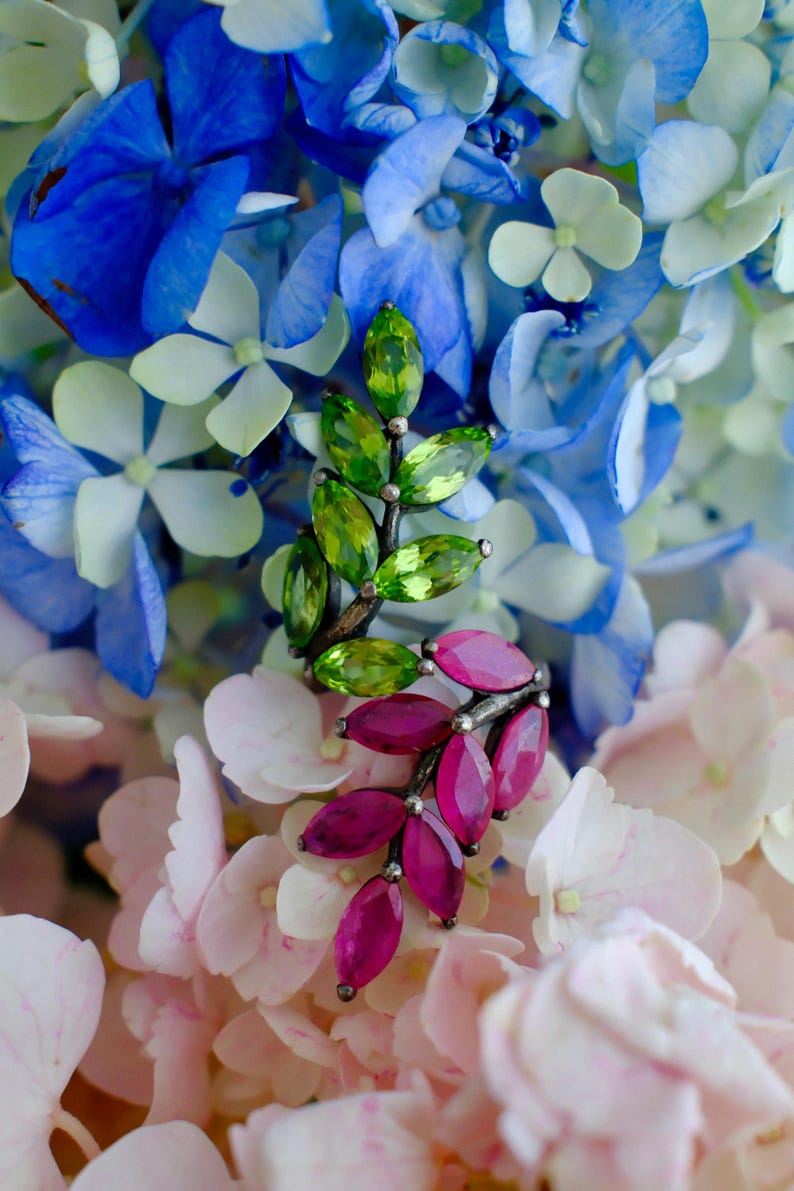 Pu&ograve; includere: Orecchini a forma di foglia con gemme verdi e magenta. Sono presentati su uno sfondo di fiori di ortensia blu, bianchi e rosa. Le gemme sono tagliate a forma ovale e disposte a forma di foglie.