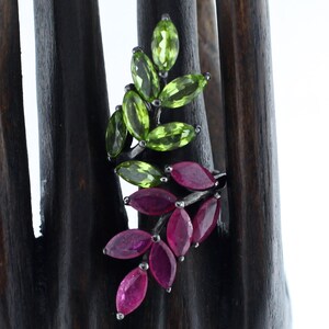 Pu&ograve; includere: Un anello d'argento con un design a foglie con pietre preziose taglio marquise. La parte superiore dell'anello presenta pietre di peridoto verde, mentre la sezione inferiore mostra rubini rossi vibranti. L'anello &egrave; esposto su un supporto di legno scuro.