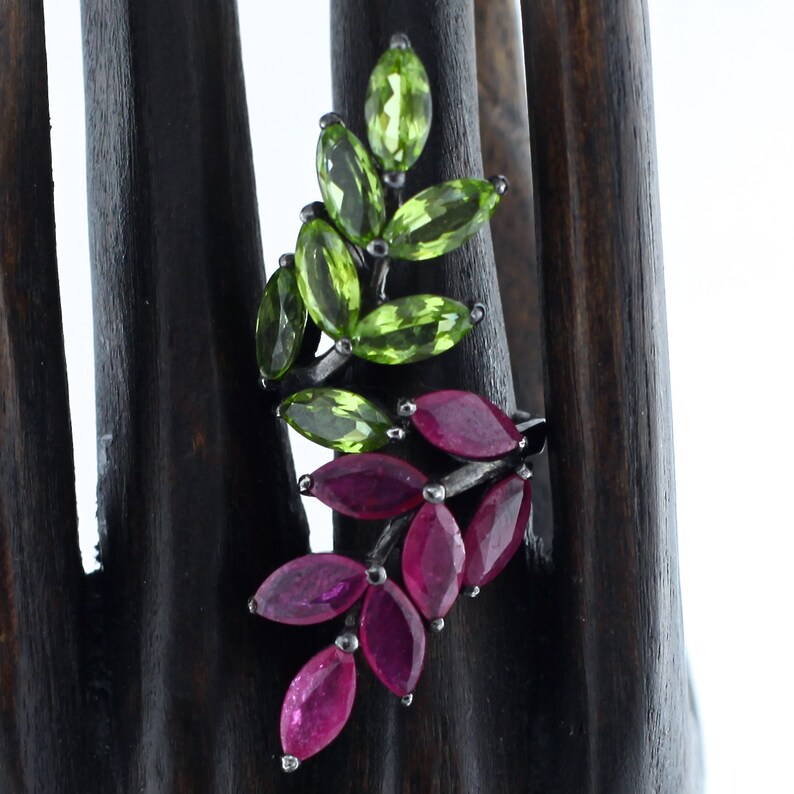 Pu&ograve; includere: Un anello d'argento con un design a foglie con pietre preziose taglio marquise. La parte superiore dell'anello presenta pietre di peridoto verde, mentre la sezione inferiore mostra rubini rossi vibranti. L'anello &egrave; esposto su un supporto di legno scuro.