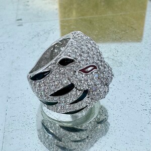 Puede incluir: Un anillo de plata con un dise&ntilde;o de tigre blanco y negro. La cara del tigre est&aacute; cubierta de peque&ntilde;as piedras transparentes. El anillo est&aacute; colocado sobre una superficie reflectante.